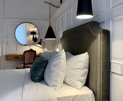 Una cama bien hecha con almohadas blancas y azul turquesa, un cabecero oscuro y un espejo redondo encima.
