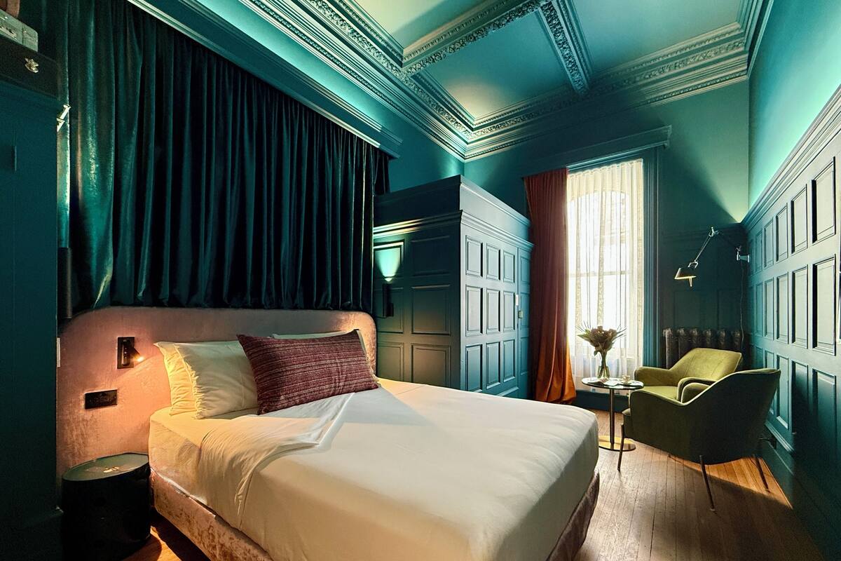 A bedroom with a large bed, a nightstand, a chair, a desk, and a window with curtains.