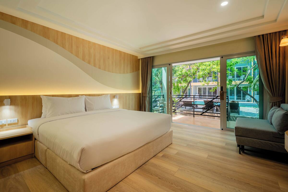 Une chambre d’hôtel moderne dotée d’un grand lit, d’un canapé et d’un balcon offrant une vue sur la verdure et une piscine.