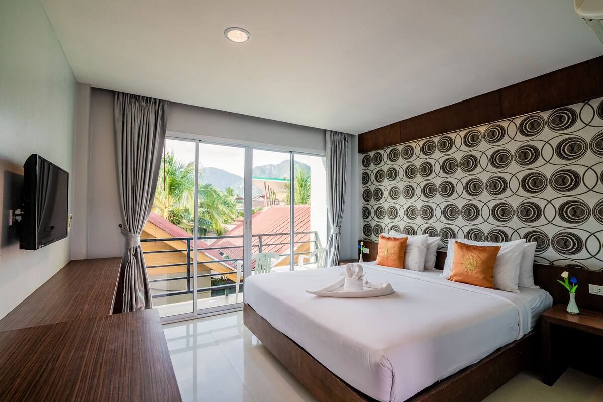 Une chambre d’hôtel avec un grand lit, une télévision, un balcon offrant une vue sur les palmiers et les immeubles, et un mur décoré de motifs.