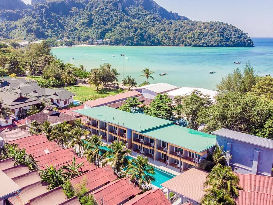 A resort with multiple buildings, a swimming pool, and a view of a turquoise sea and a forested hill.