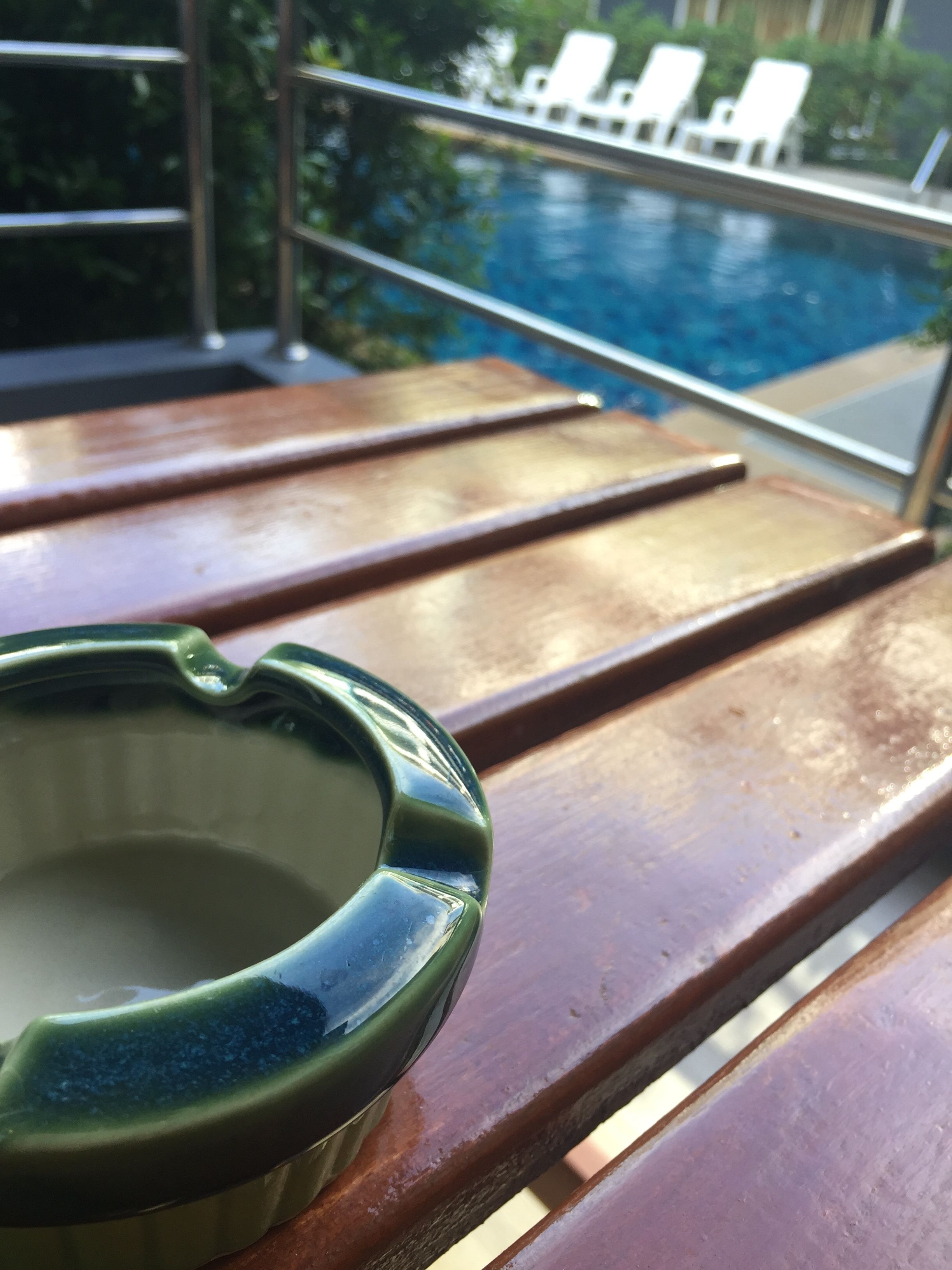 A wooden lounge chair with a green ashtray and a pool in the background.