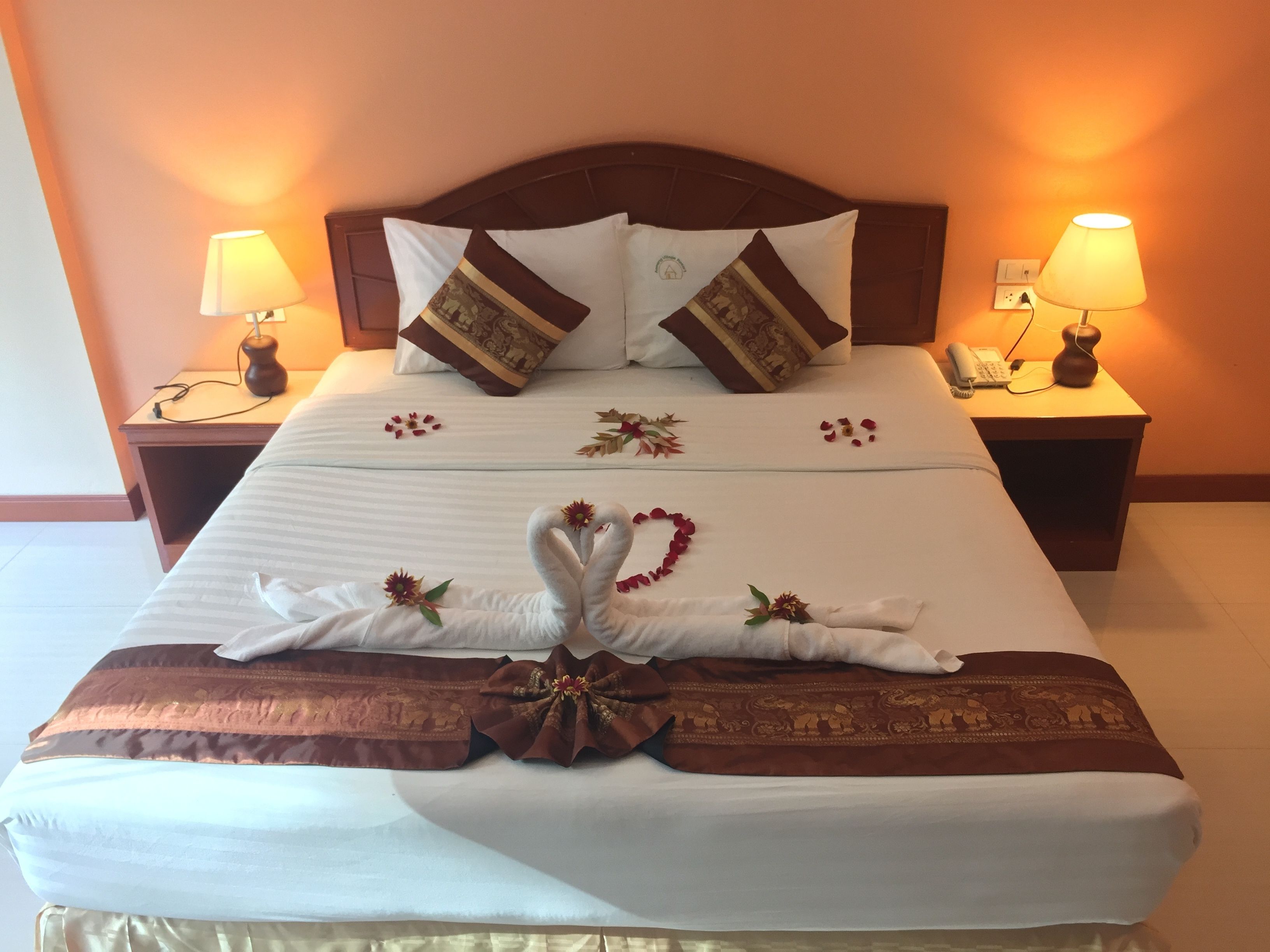 Una cama bien tendida con cisnes decorativos y flores, dos lámparas de noche y un teléfono en la mesita.