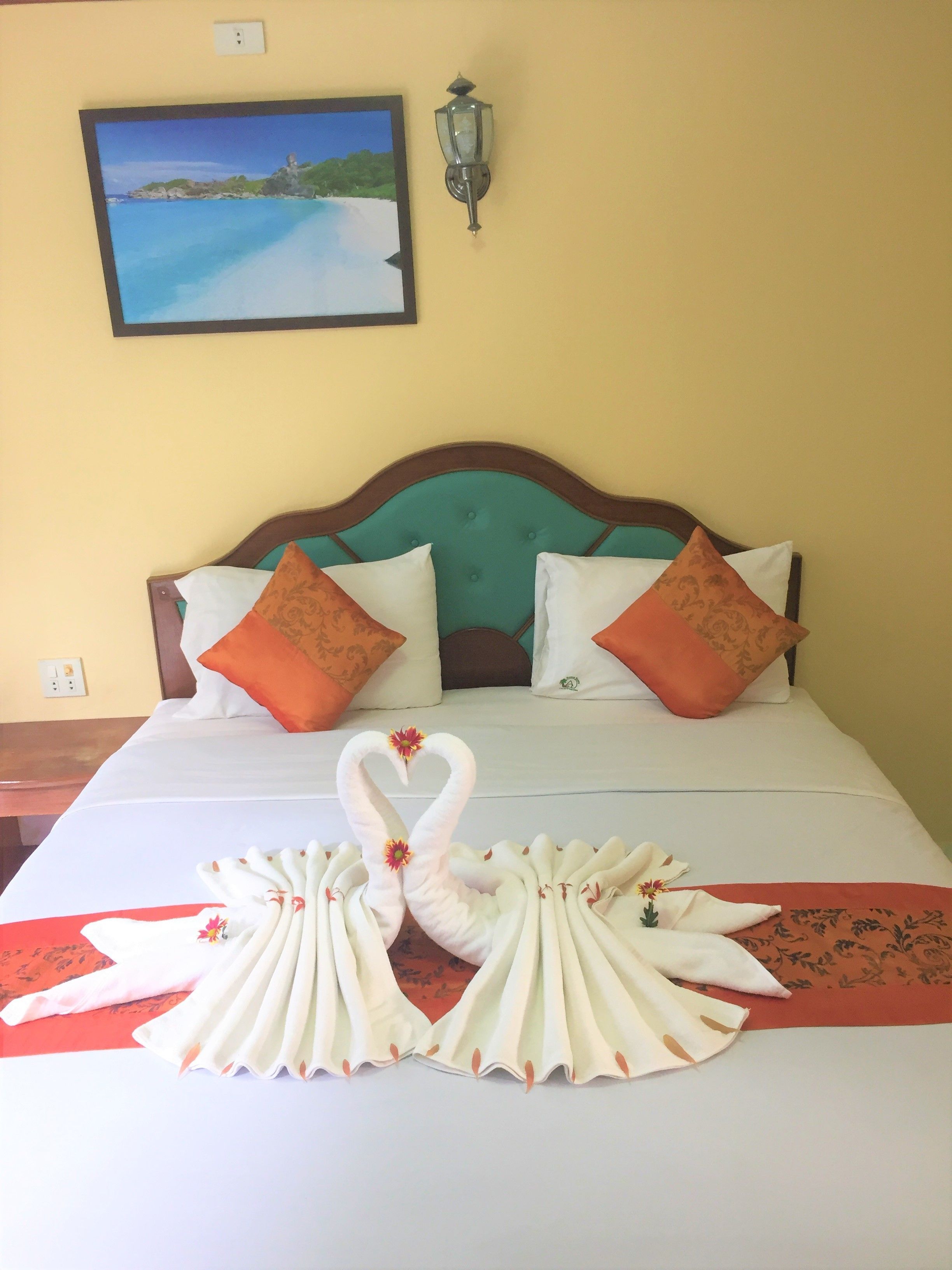 Habitación de hotel con cama, almohadas, una lámpara de pared y un cuadro de una playa.