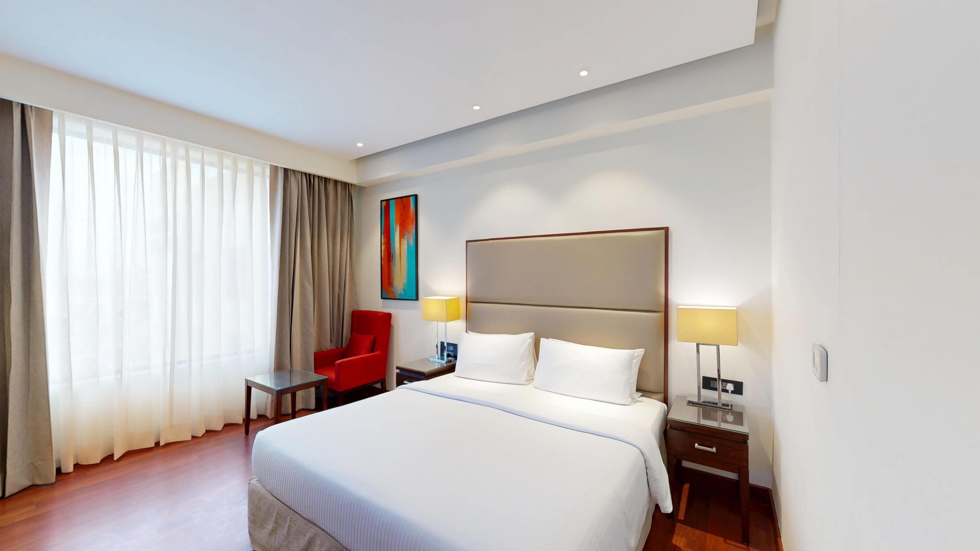 A hotel room with a large bed, a red armchair, two bedside tables with lamps, and a window with sheer curtains.