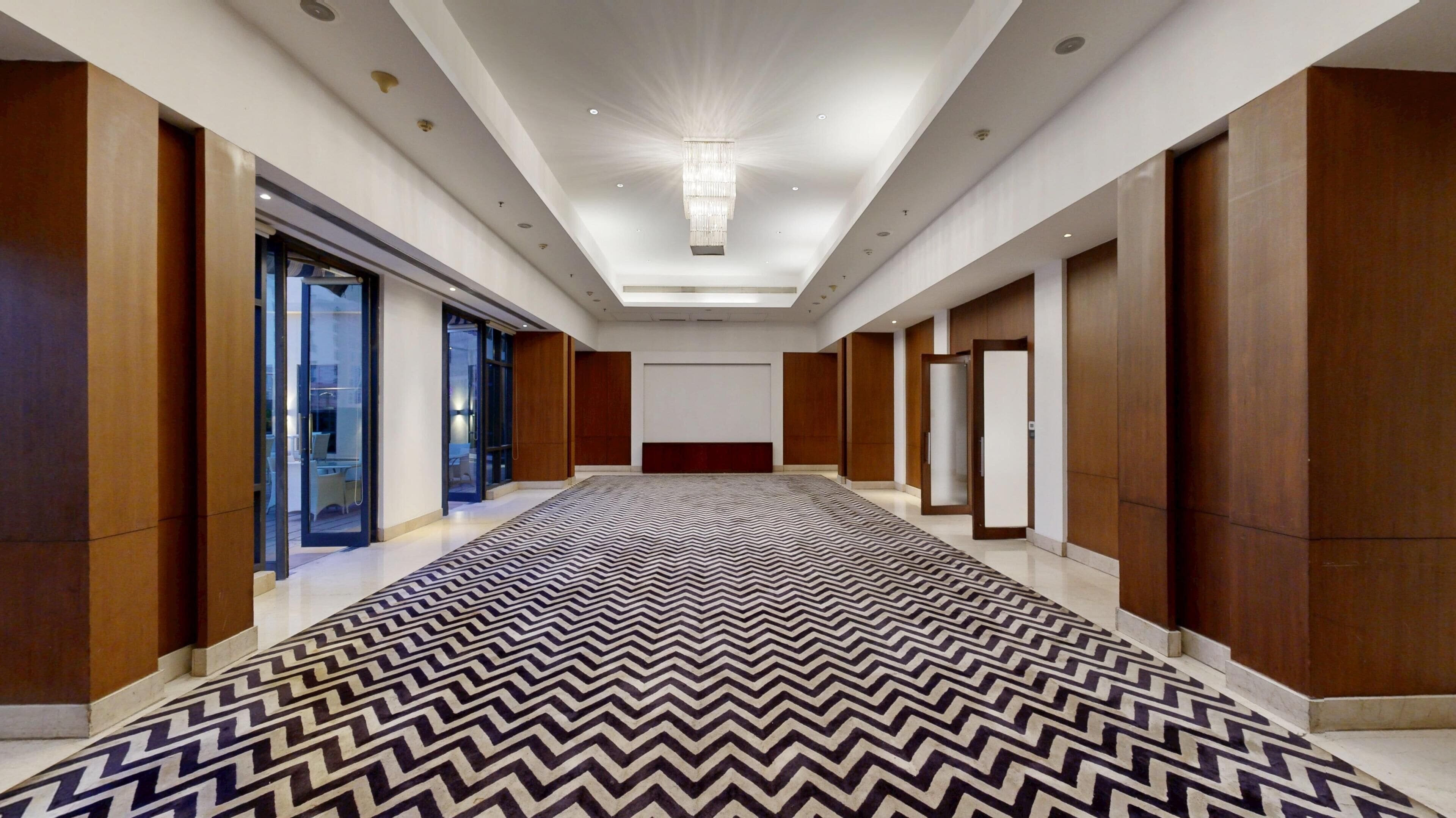 A hotel corridor with a patterned carpet, wooden panel walls, and a central chandelier.