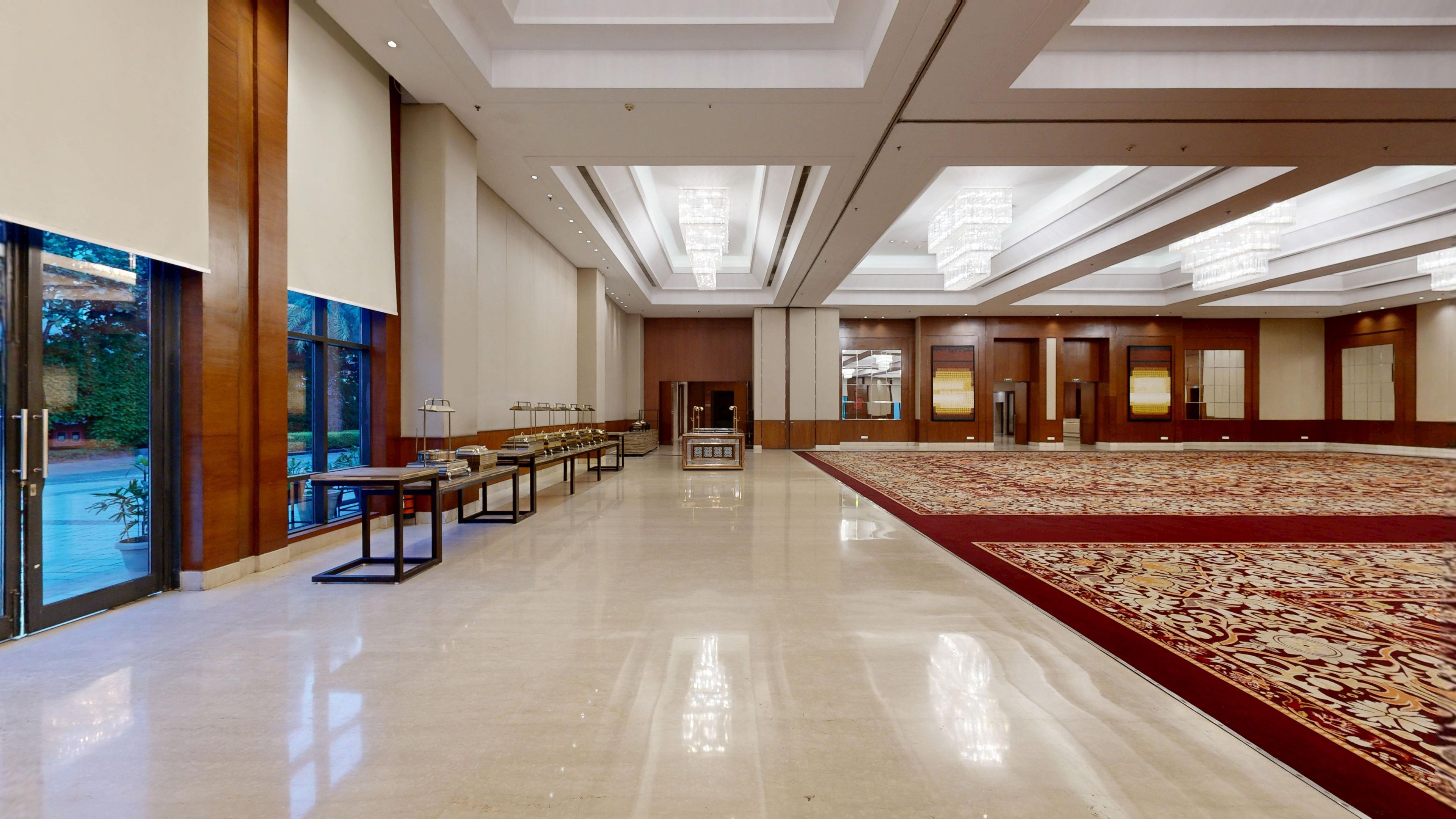 A spacious hotel lobby with a long reception desk, a large carpeted area, and a view through the glass doors.