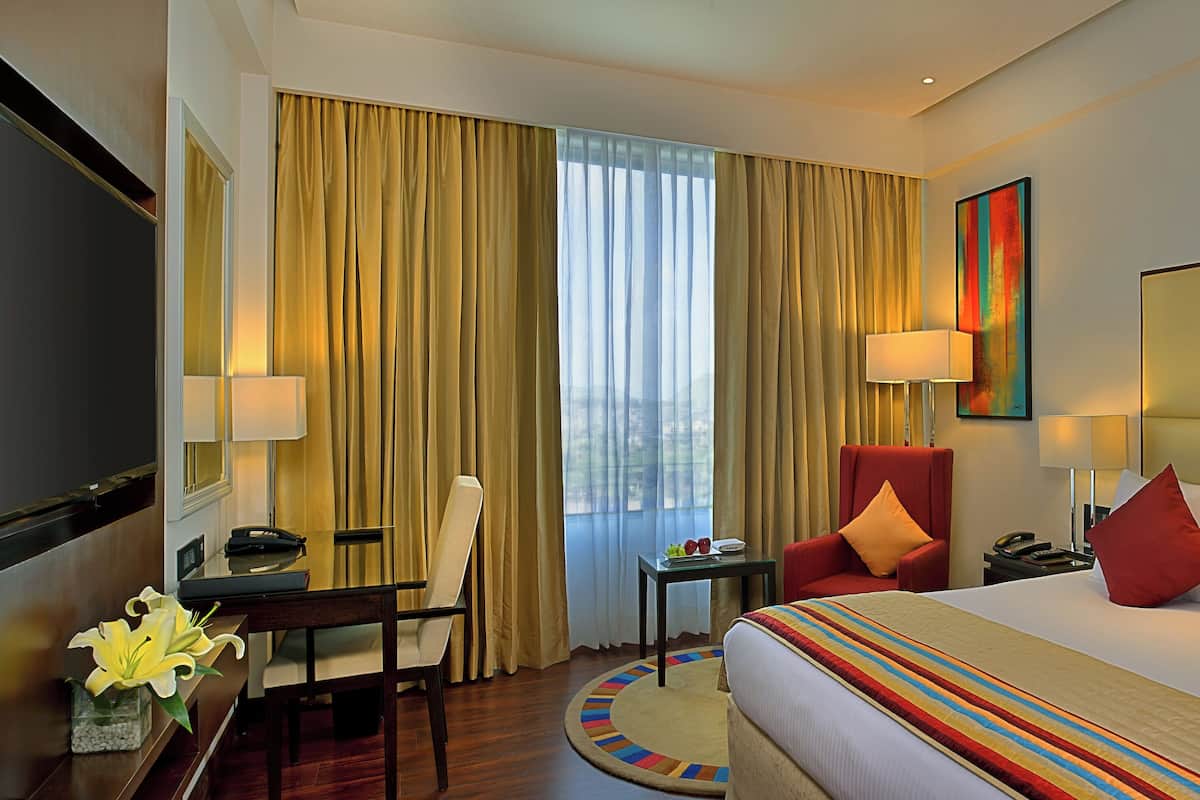 Une chambre d’hôtel comprenant un lit, un bureau avec un téléphone, une chaise, une table avec une lampe et une fenêtre avec des rideaux.