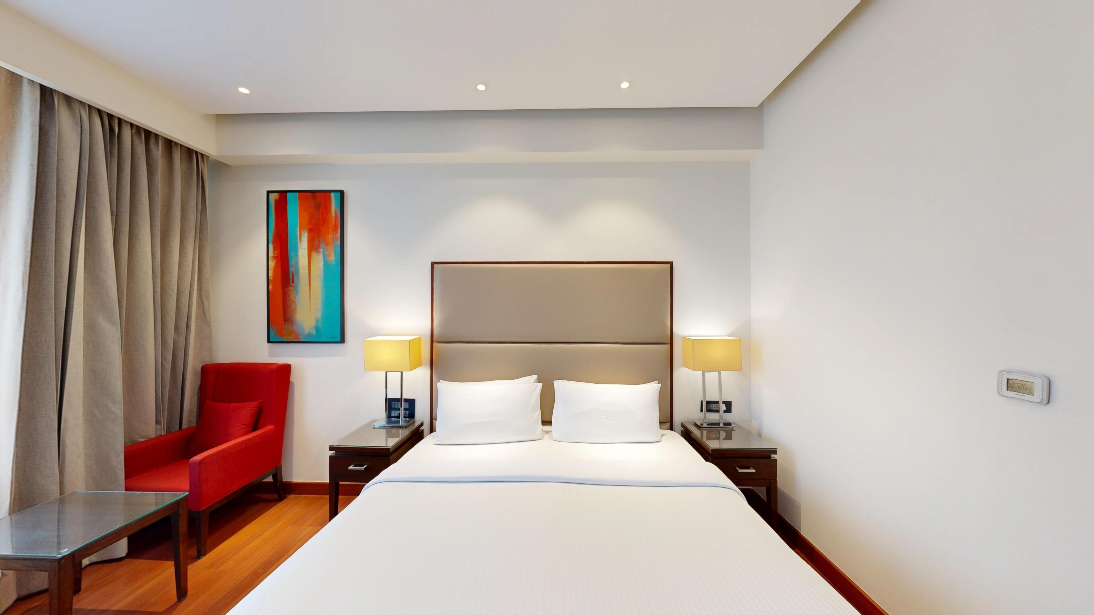 A hotel room with a large bed, two bedside tables with lamps, a red armchair, a painting on the wall, and a window with curtains.