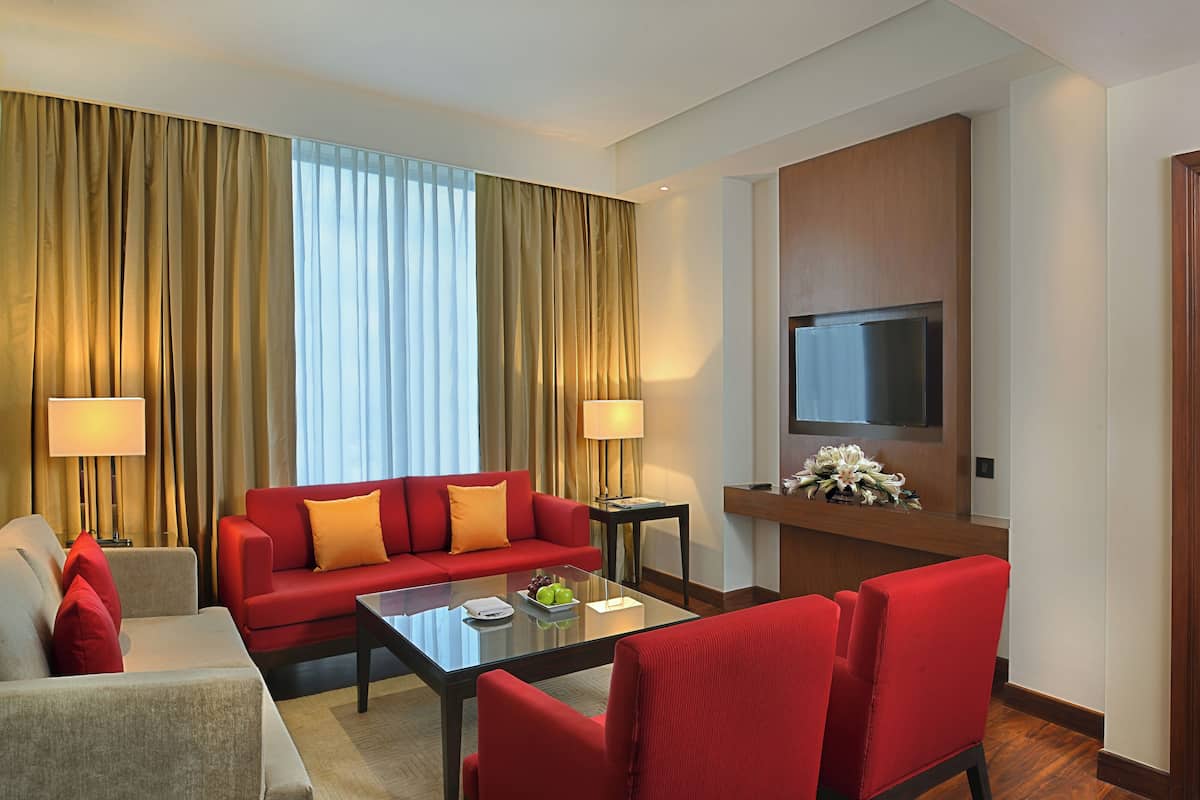 Une chambre d’hôtel avec un canapé rouge, une table basse en verre, un téléviseur et un bureau avec une lampe.