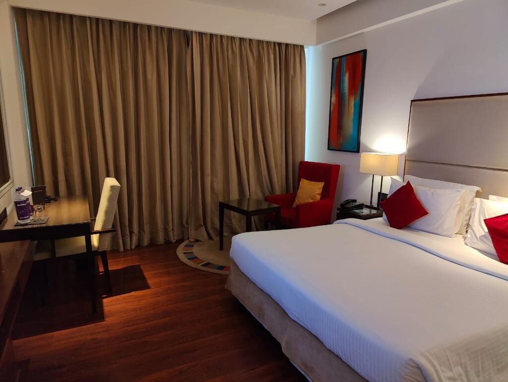 A hotel room with a bed, a desk, a chair, a lamp, and a painting on the wall.