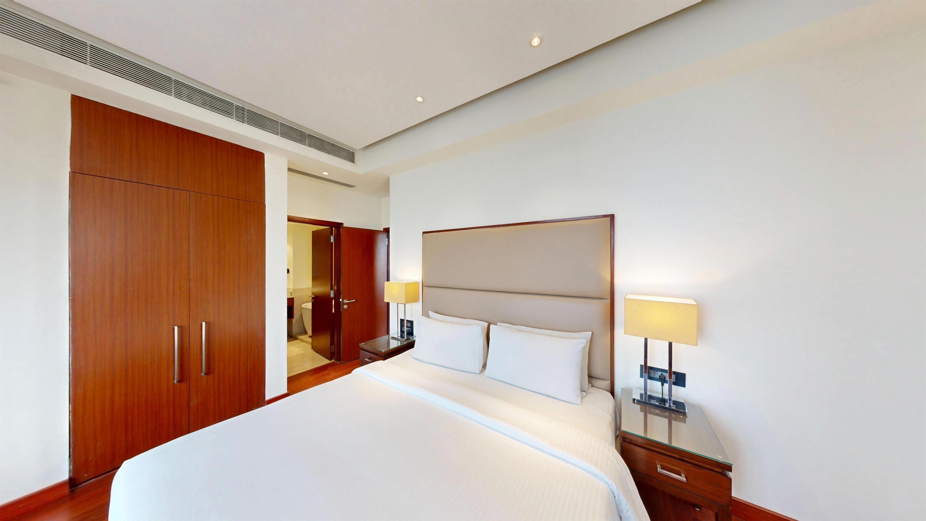 Habitación de hotel con una cama grande, un armario de madera, lámparas de noche y un baño visible a través de una puerta abierta.