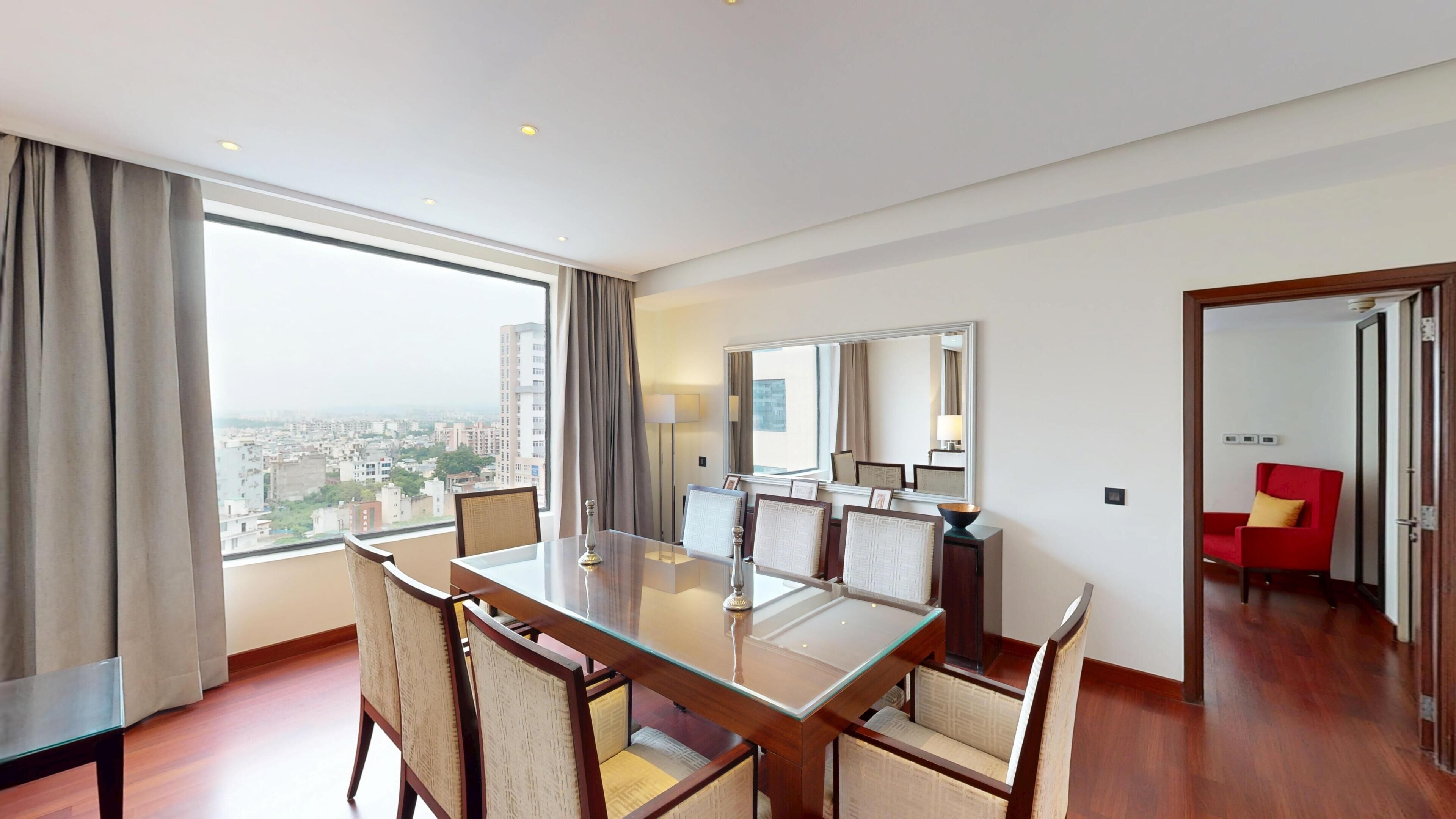 Habitación de hotel con comedor, un ventanal con vista a la ciudad, una silla roja y un espejo en la pared.