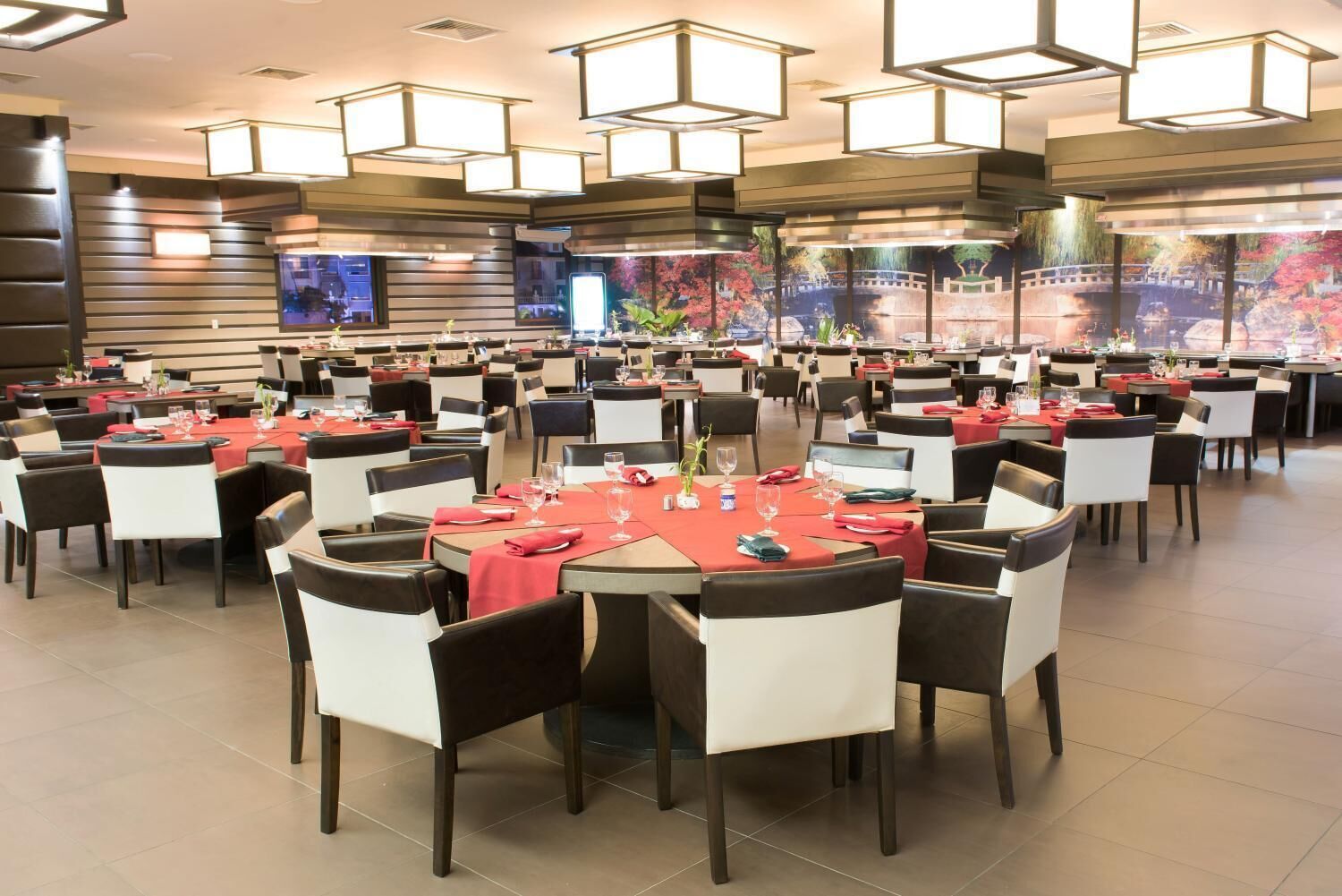 A spacious dining area with multiple tables and chairs, featuring a modern design with geometric light fixtures and a large mural on the back wall.