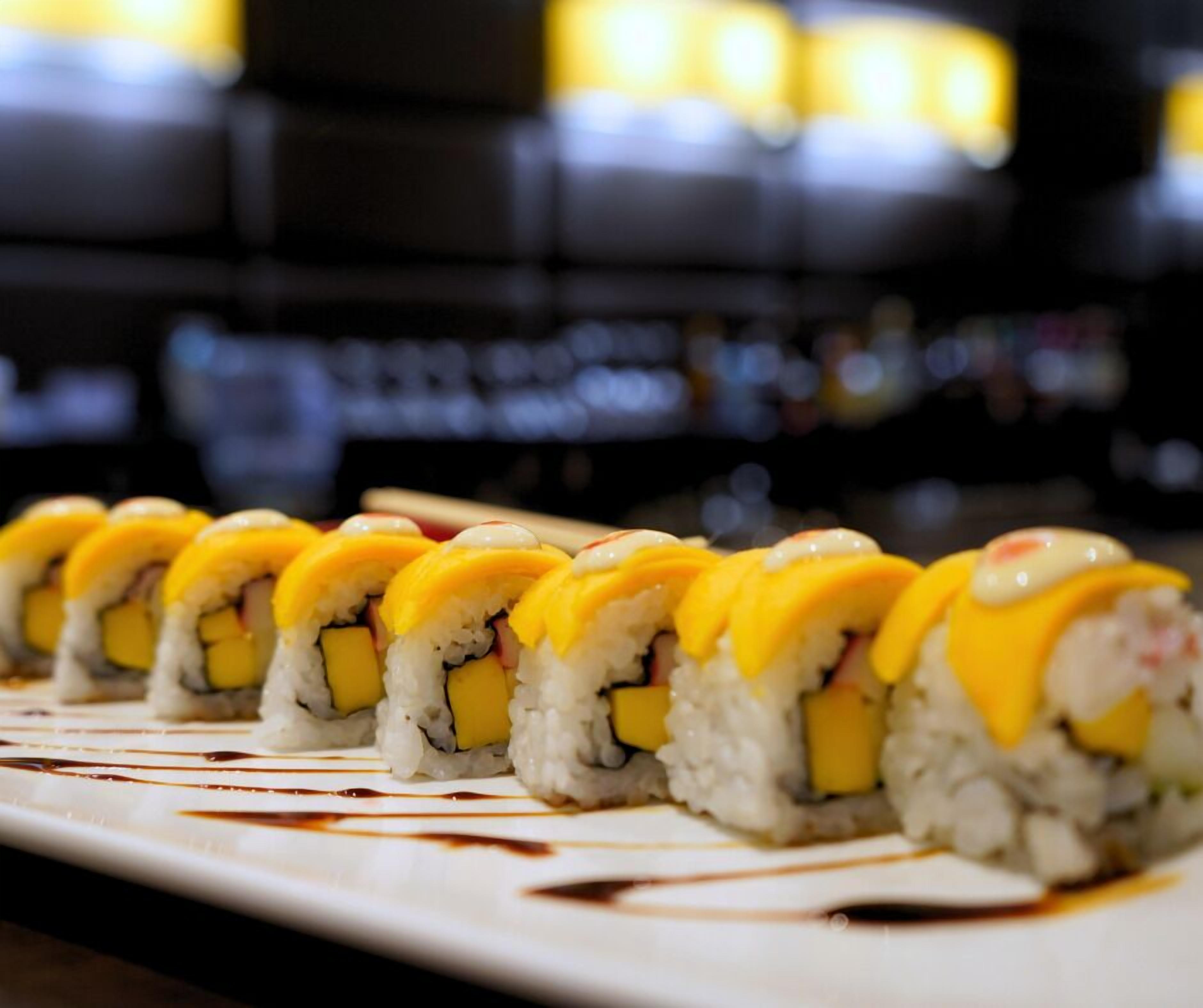 Una hilera de rollos de sushi con cobertura amarilla, dispuestos sobre un plato blanco con salpicaduras de salsa.
