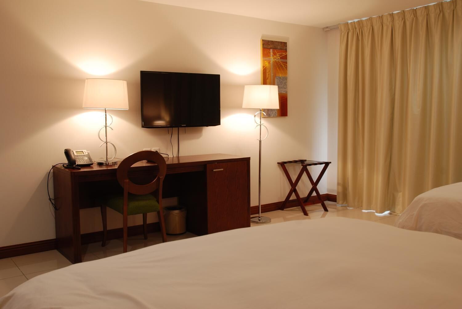 Habitación de hotel con cama, escritorio, silla, televisor, dos lámparas y ventana con cortinas.