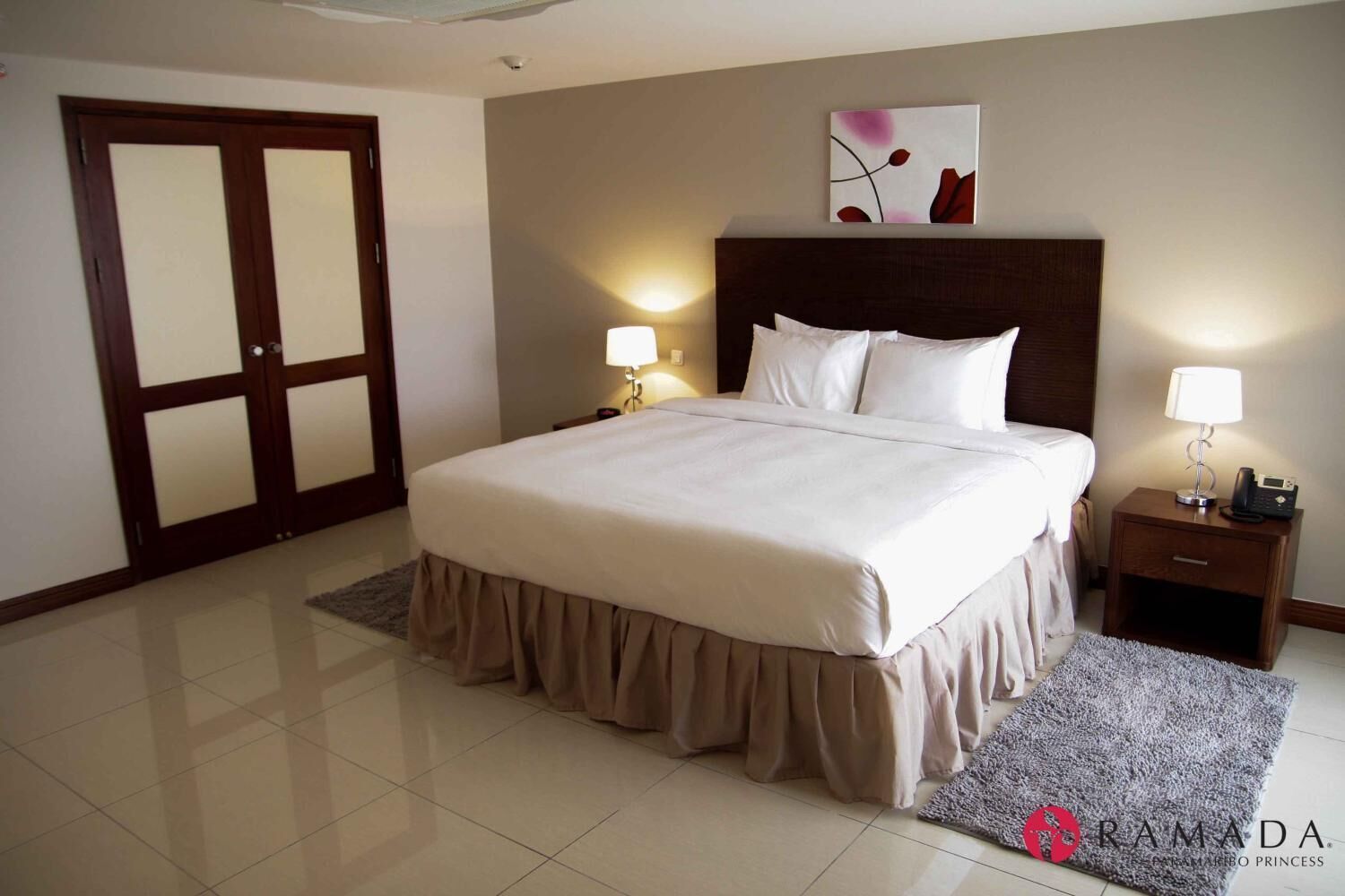 A hotel room with a large bed, two bedside lamps, a nightstand, a door with frosted glass panels, and a painting on the wall.