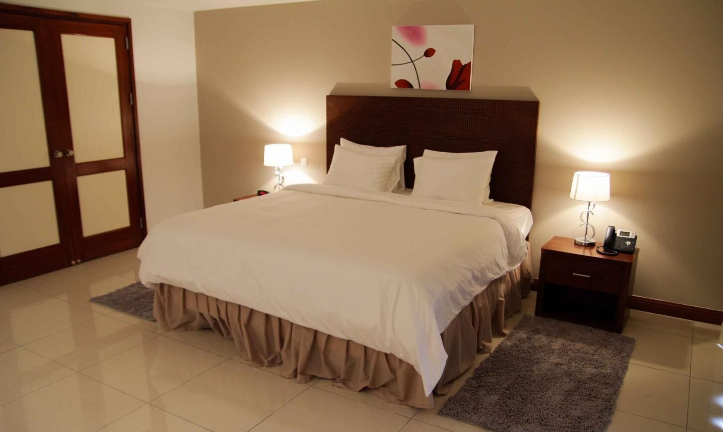 Una habitación de hotel con una cama grande, dos mesitas de noche con lámparas, un cabecero de madera y una puerta con paneles de vidrio.
