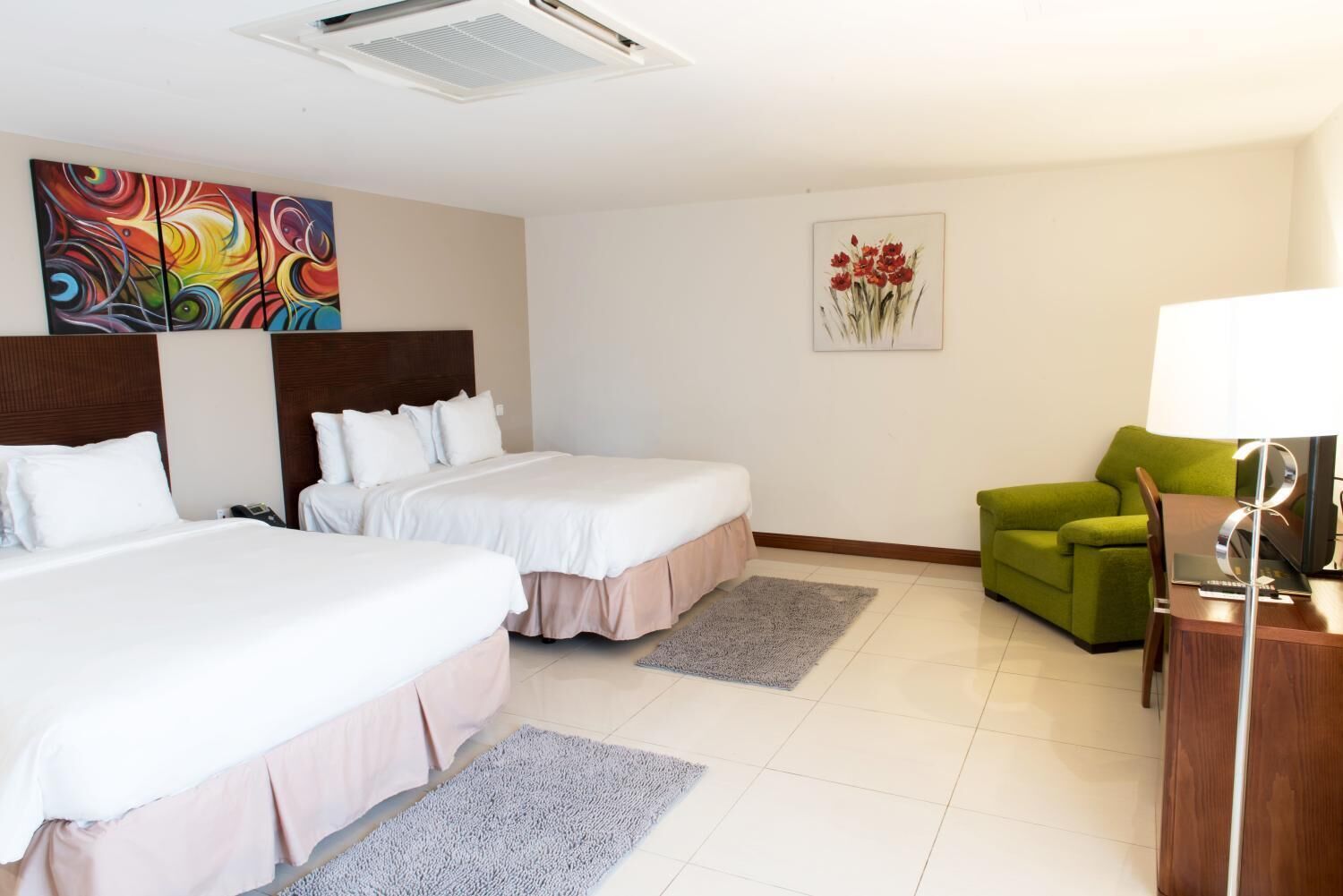 A hotel room with two beds, a green armchair, a desk with a lamp, and artwork on the walls.