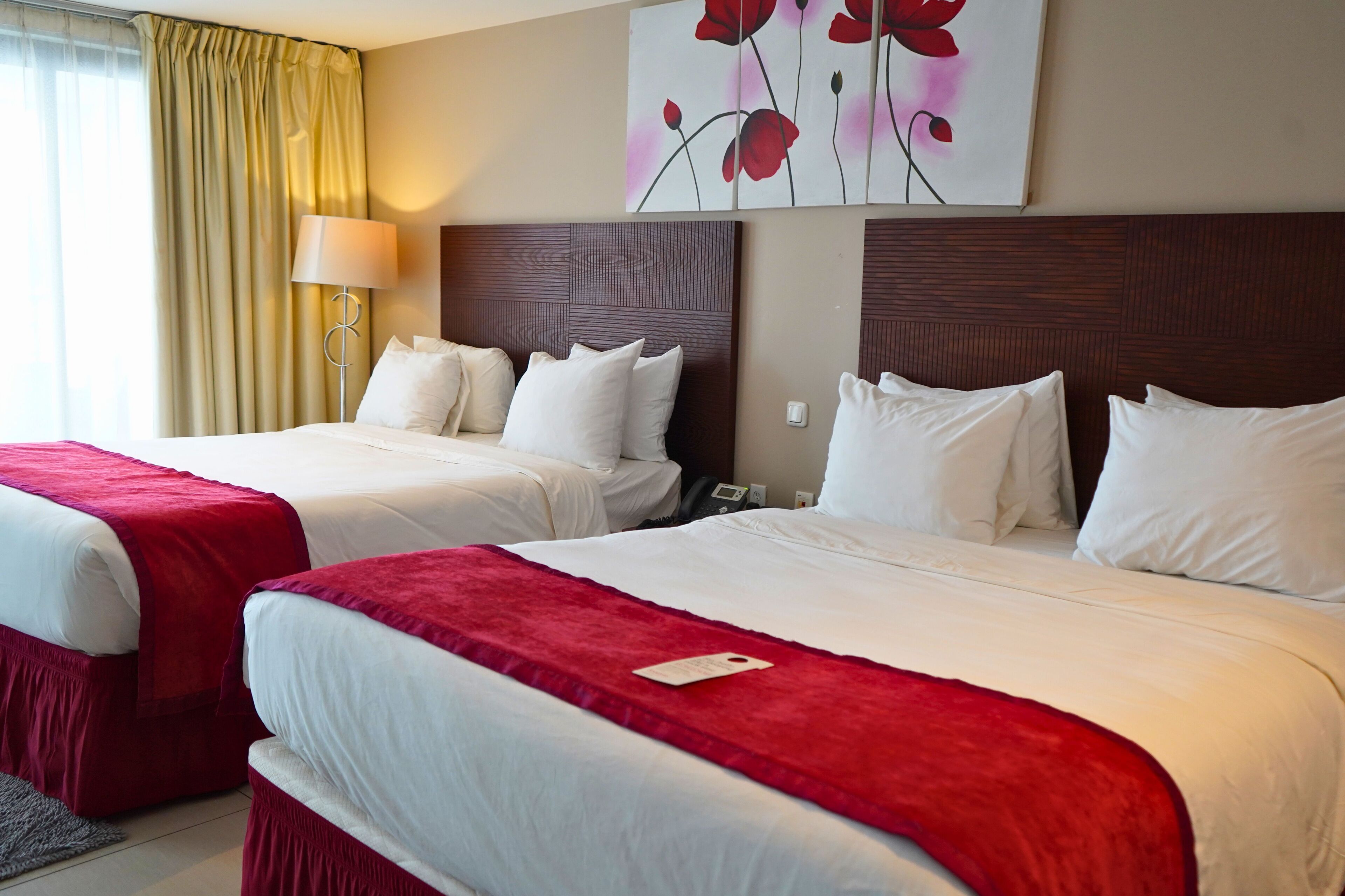 Habitación de hotel con dos camas, cada una con ropa blanca y cabeceros rojos. En la pared sobre las camas hay un cuadro de flores rojas.
