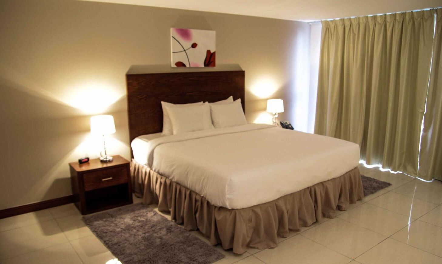 A neatly made bed with a headboard, bedside lamps, a small bedside table, and a window with curtains.