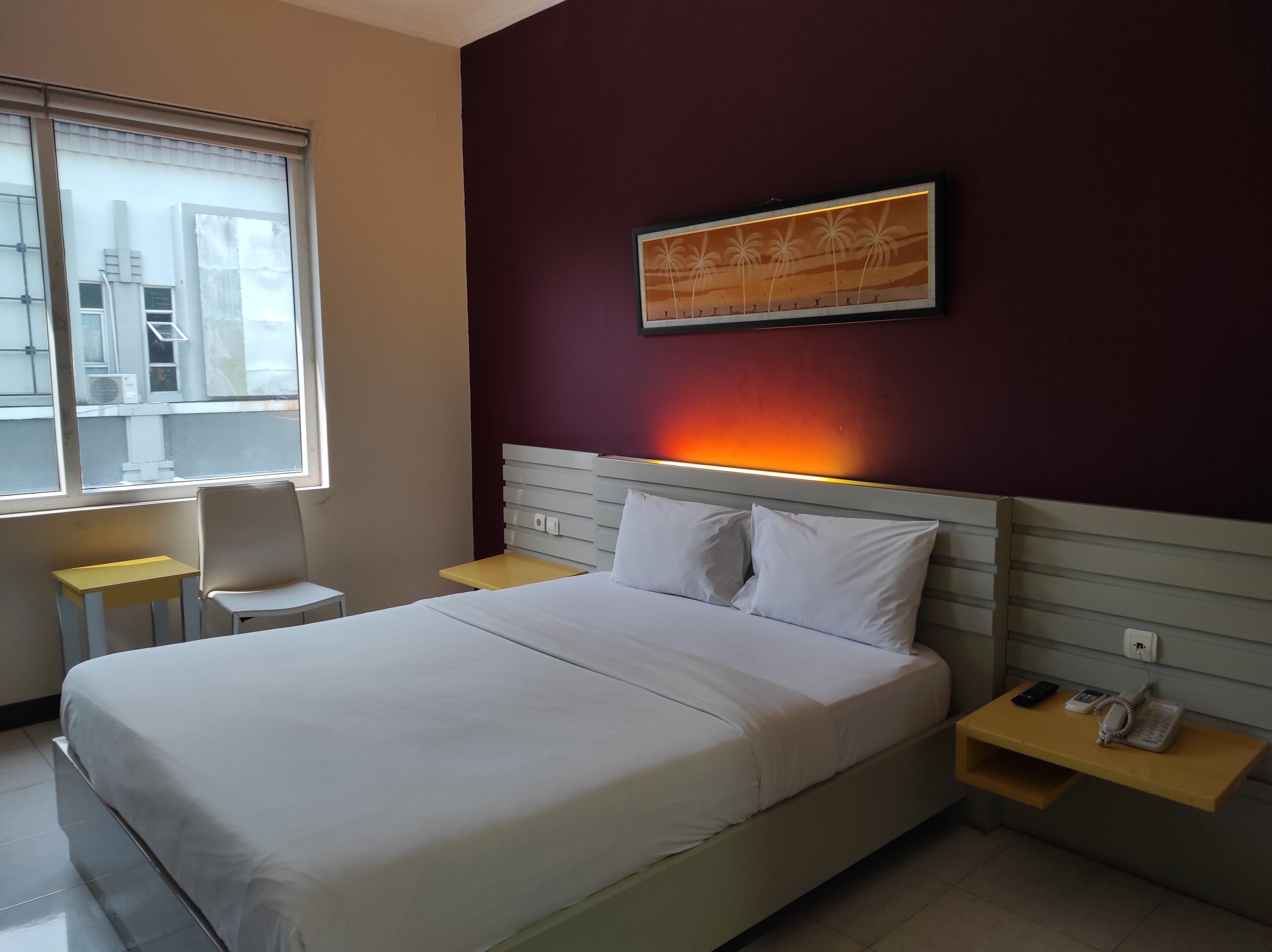 A hotel room with a bed, a chair, a small table, a bedside table with a phone, and a wall-mounted artwork.
