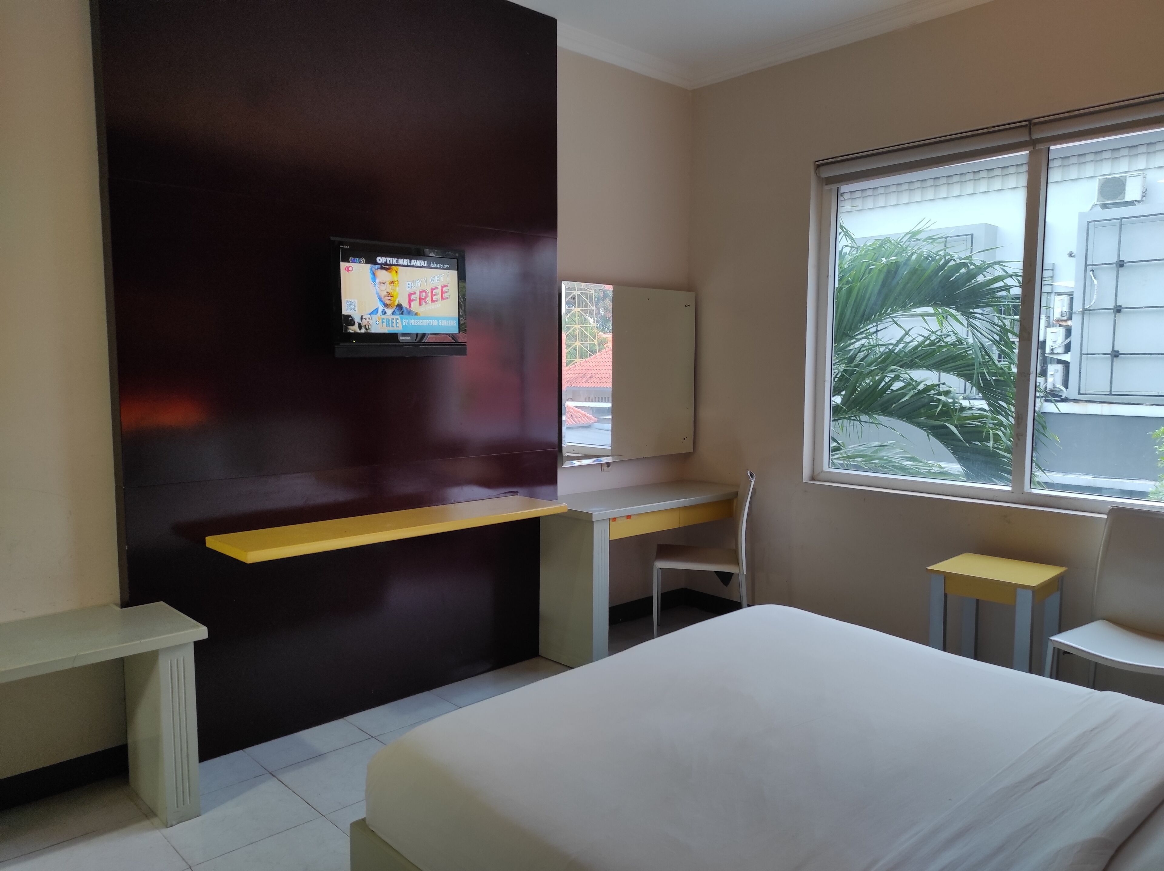 A hotel room with a bed, a desk, a chair, a TV mounted on the wall, and a window with a view of the outside.