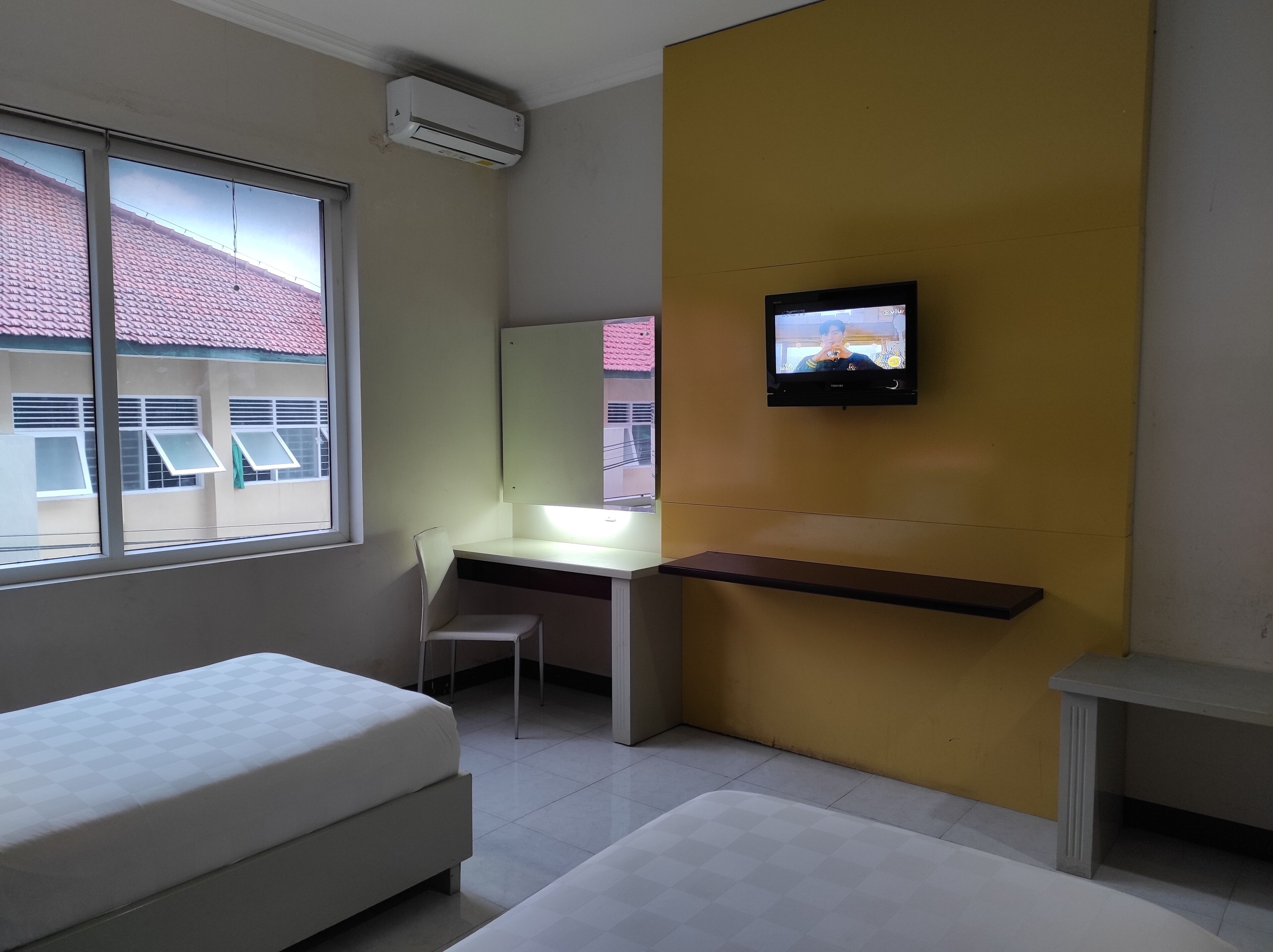 Une chambre d’hôtel moderne équipée d’un lit, d’un bureau, d’une chaise, d’un téléviseur fixé au mur et d’une fenêtre donnant sur des immeubles.