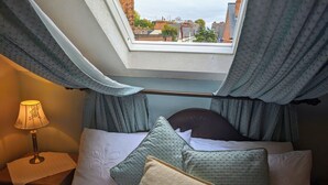 A bedroom with a skylight, a bed with multiple pillows, a bedside table with a lamp, and a view of buildings through the window. - The Sycamore Guest House (York)