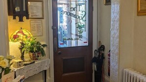 A hallway with a wooden door, a clock, a chandelier, a radiator, and a table with a lamp and flowers. - The Sycamore Guest House (York)