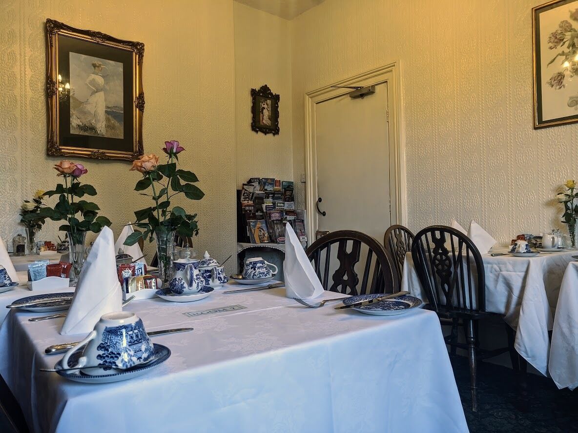 A dining area with tables set for a meal, featuring blue and white porcelain dishes, a vase with roses, and framed artwork on the walls.
