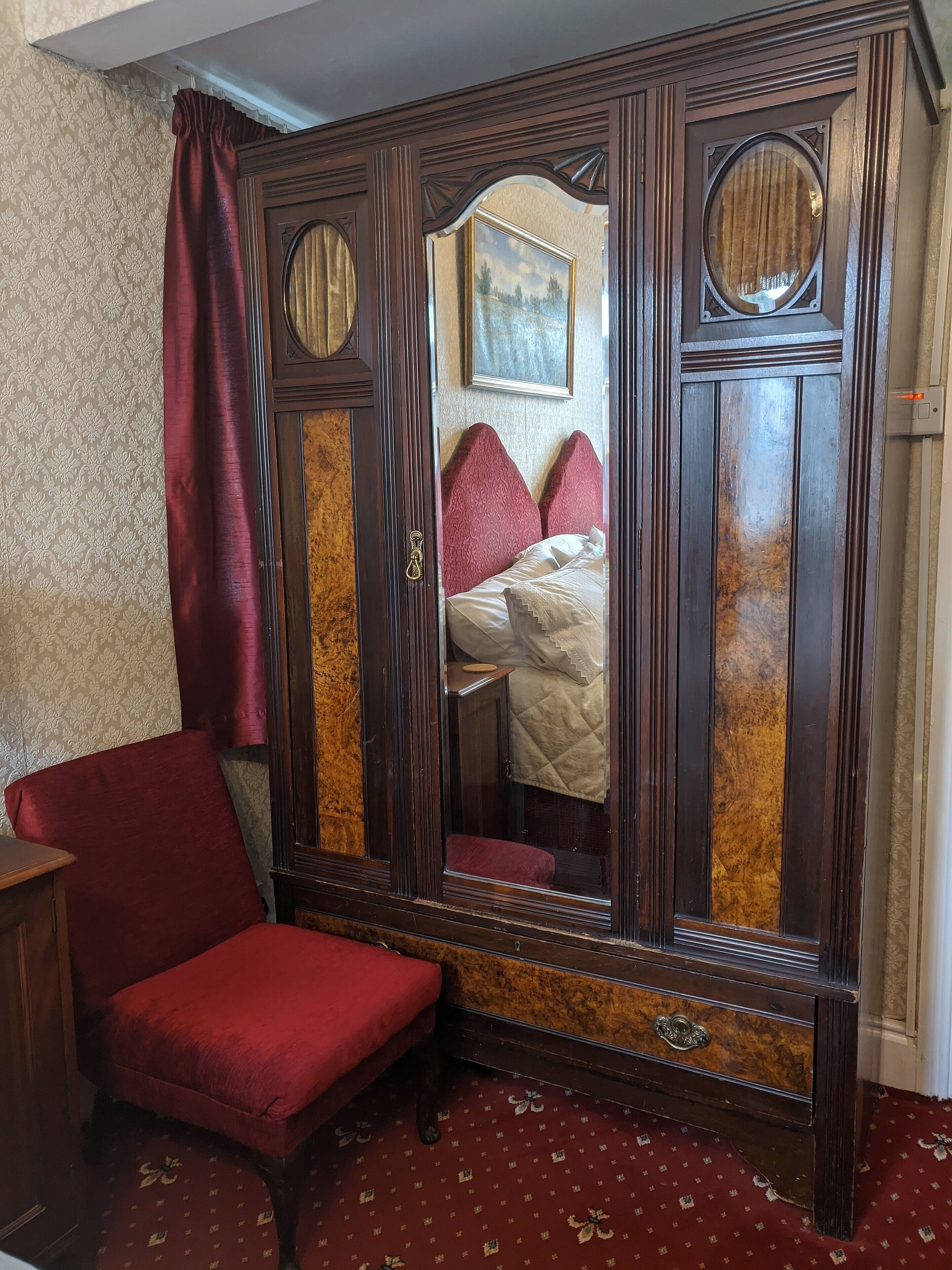 A room with a large mirror, a red chair, a bed with red pillows, and a patterned carpet.