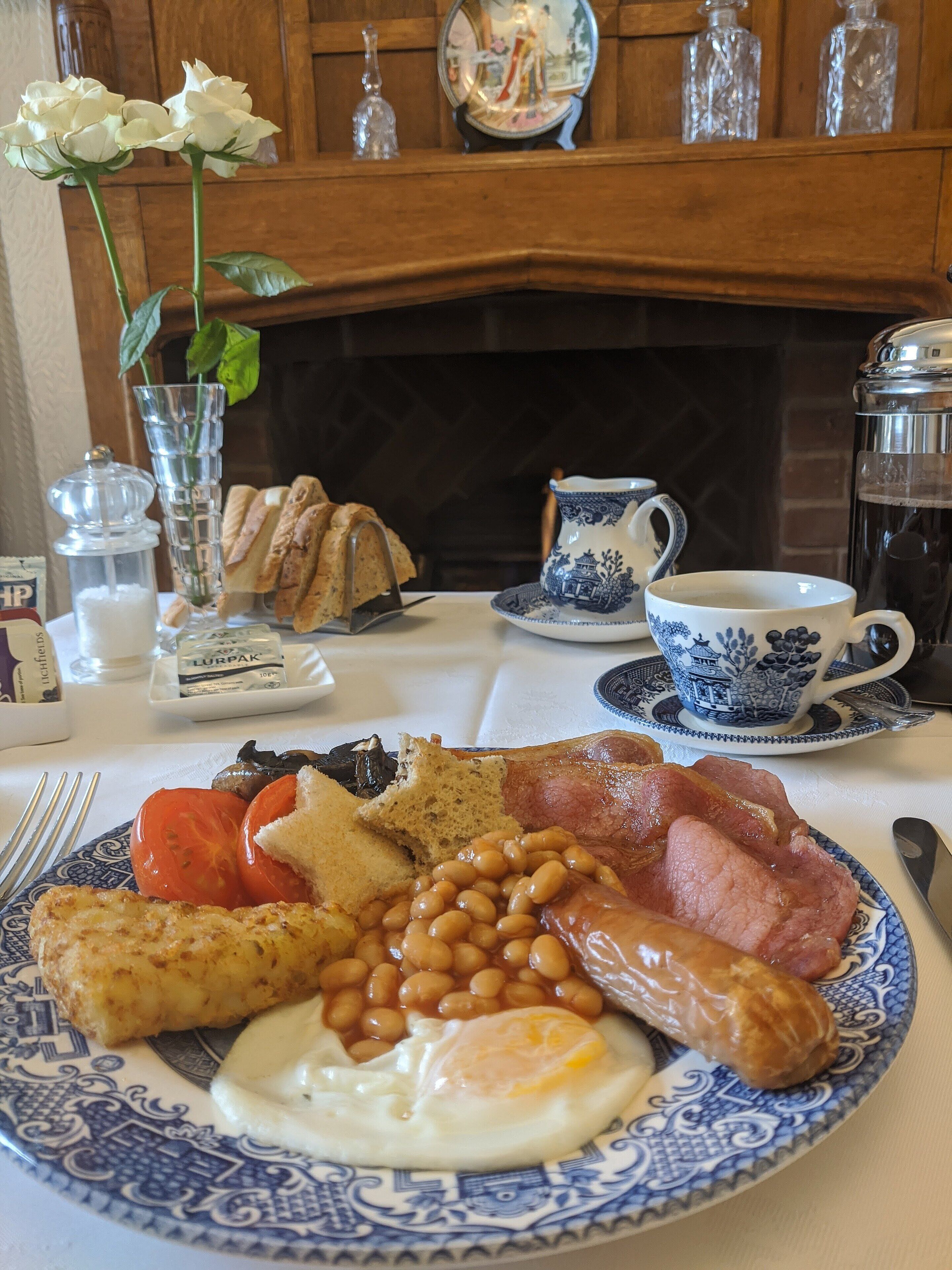 A breakfast table with a plate of eggs, beans, bacon, and toast, a teapot, and a vase with flowers.