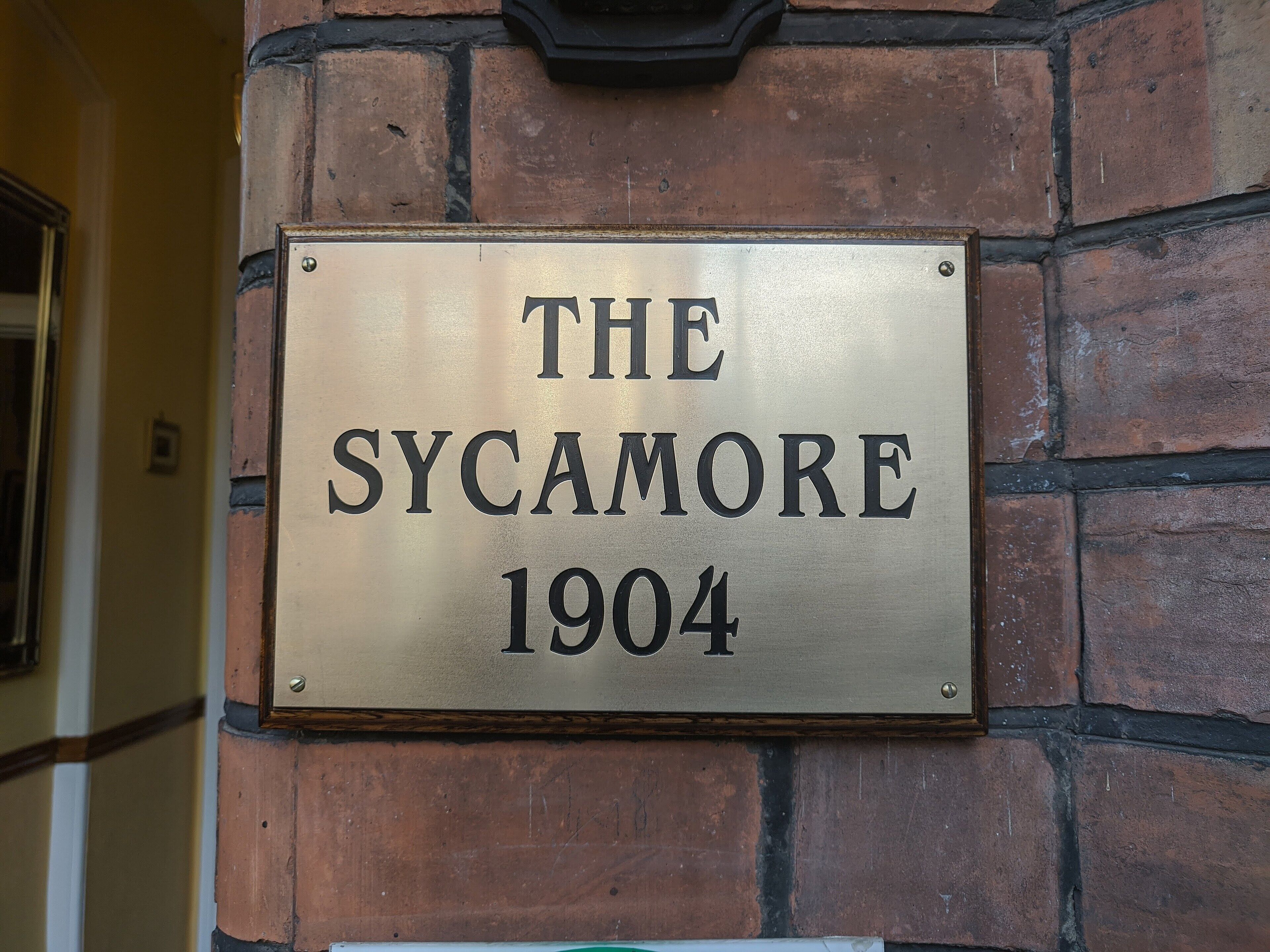 Une plaque en métal fixée à un mur de briques, portant l’inscription : « THE SYCAMORE 1904 ».