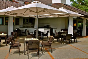 An outdoor patio area with wicker chairs and tables, a large umbrella, and a kitchen area in the background. - Golf Hotel La Pinetina (Carbonate)