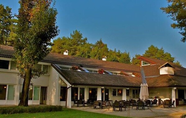 A two-story building with a sloped roof, outdoor seating area with tables and chairs, and a large tree in the foreground. - Golf Hotel La Pinetina (Carbonate)