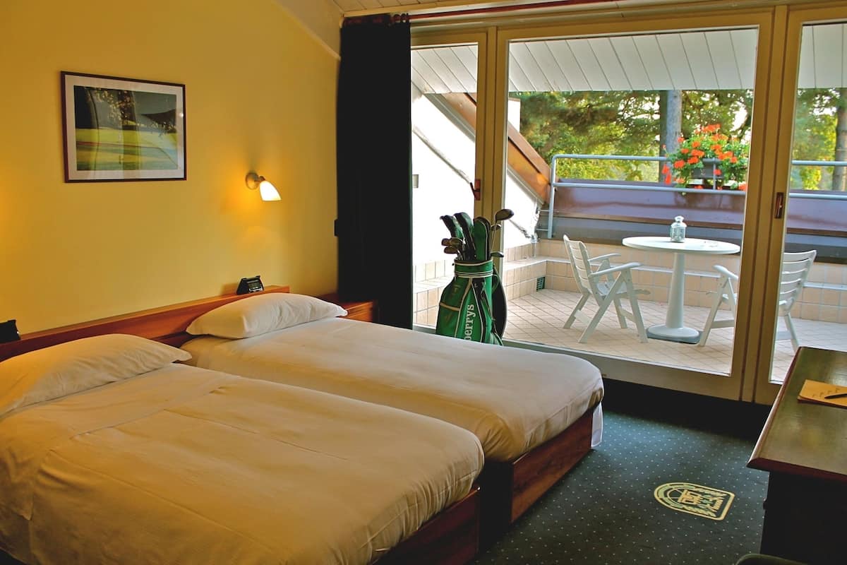 A hotel room with two beds, a balcony with a table and chairs, and a view of greenery outside.