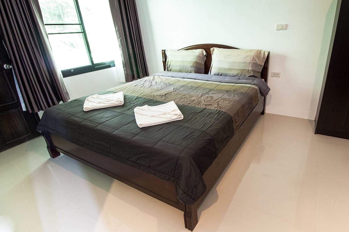 A neatly made bed with a dark bedspread and two folded towels, a wooden headboard, a window with curtains, and a dark wardrobe with two drawers.