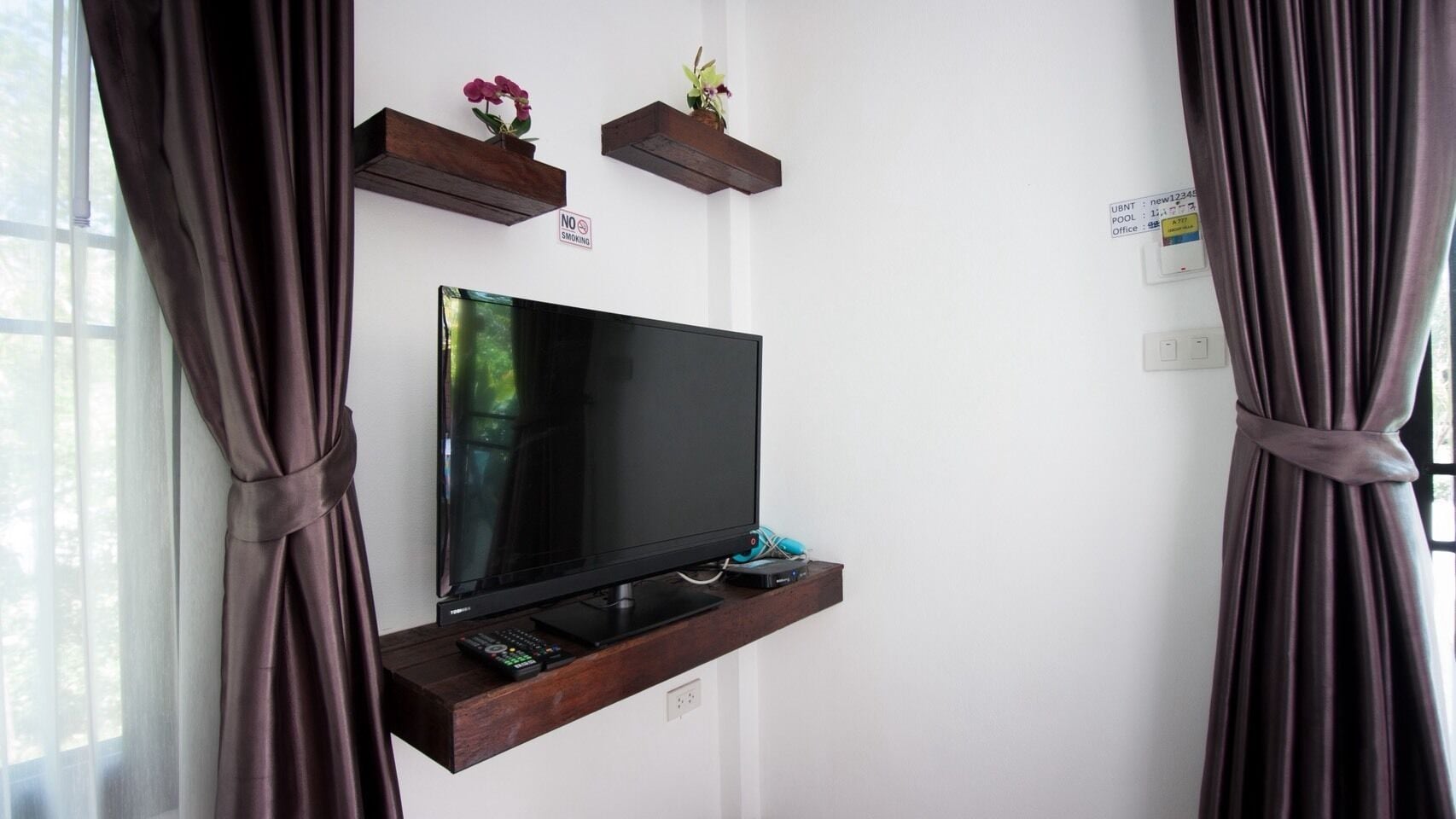 Un téléviseur posé sur une étagère en bois, avec une télécommande, un panneau indiquant «Il est interdit de fumer» et une prise électrique fixée au mur.