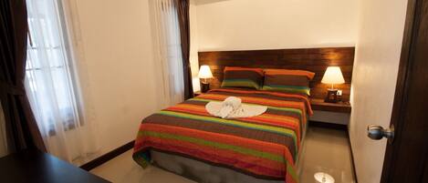 A hotel room with a bed, two bedside lamps, a wooden headboard, and a window with curtains.