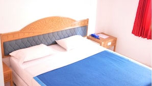 A neatly made bed with a blue bedspread, a wooden headboard, and a nightstand with a lamp and a blue item on it. - Highfive Guest House (Pattaya)
