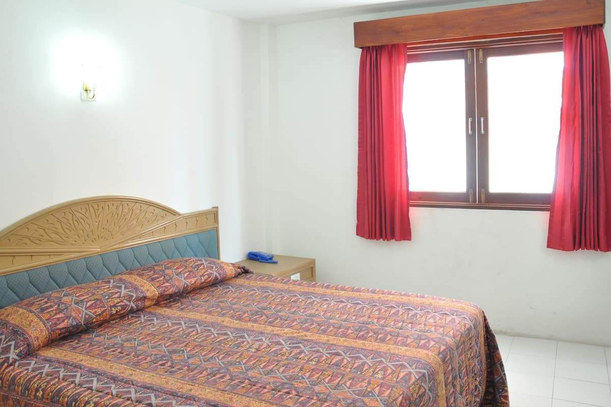 A bedroom with a bed, a nightstand, a window with red curtains, and a wall-mounted light.