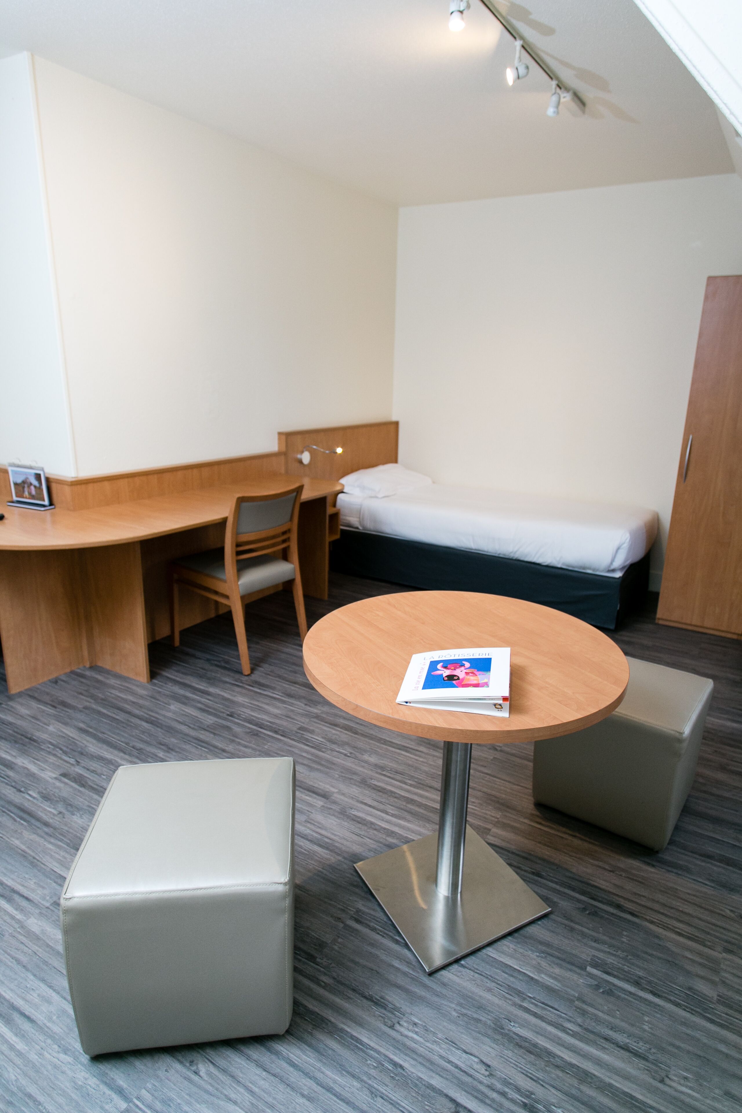 A compact hotel room with a bed, a desk, a chair, a small table, and two ottomans.
