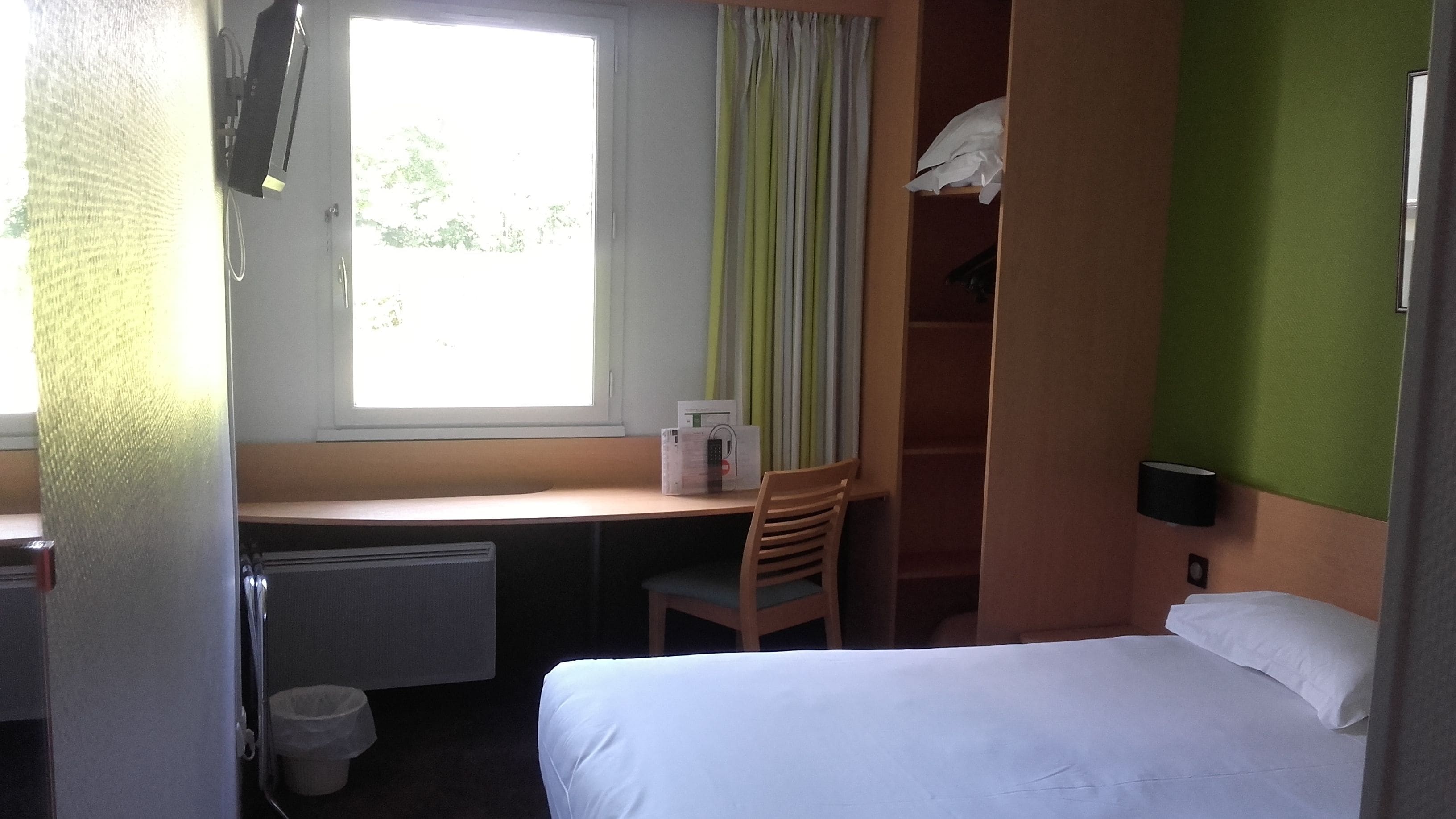 Quarto de hotel com cama, escrivaninha com cadeira, radiador, janela com cortinas e parede verde.