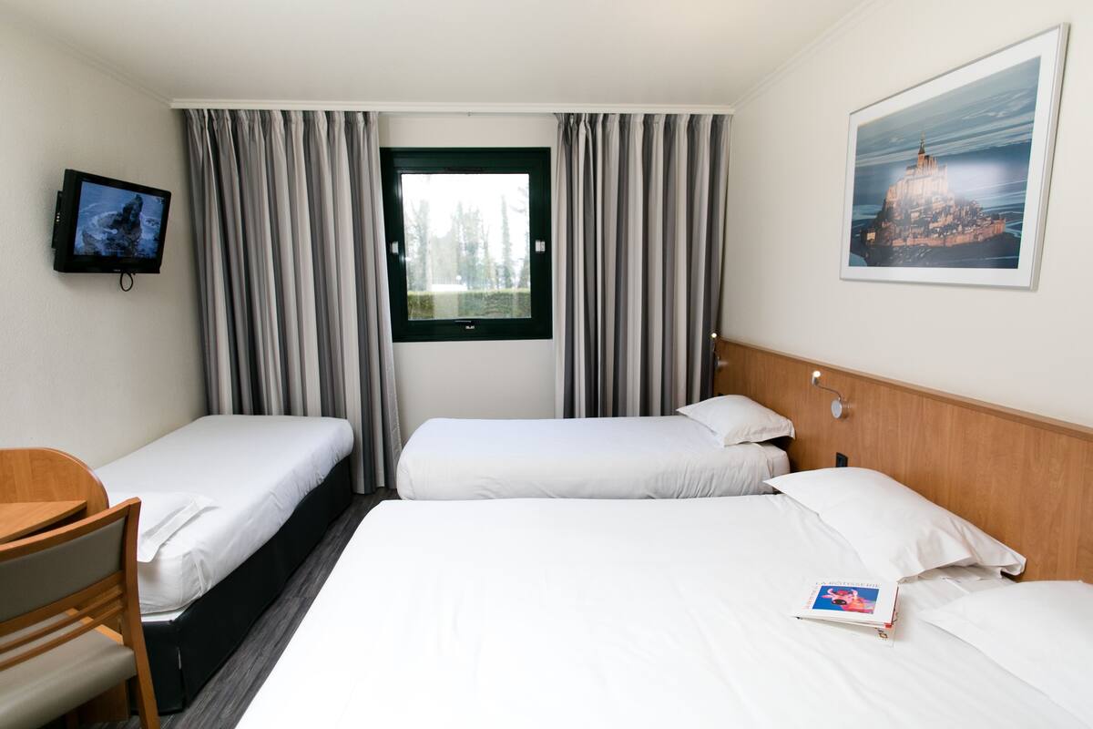 A hotel room with two single beds, a desk, a TV, and a window with curtains.