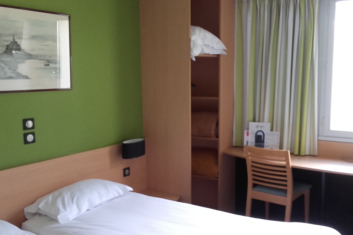 A hotel room with a bed, a desk with a chair, a window with curtains, and a wall-mounted picture.