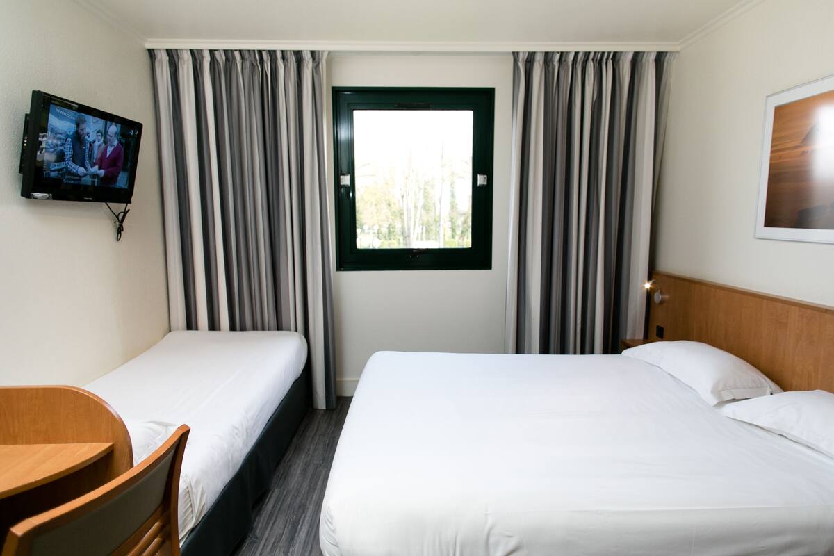 A hotel room with two single beds, a TV mounted on the wall, a window with curtains, and a wooden headboard.