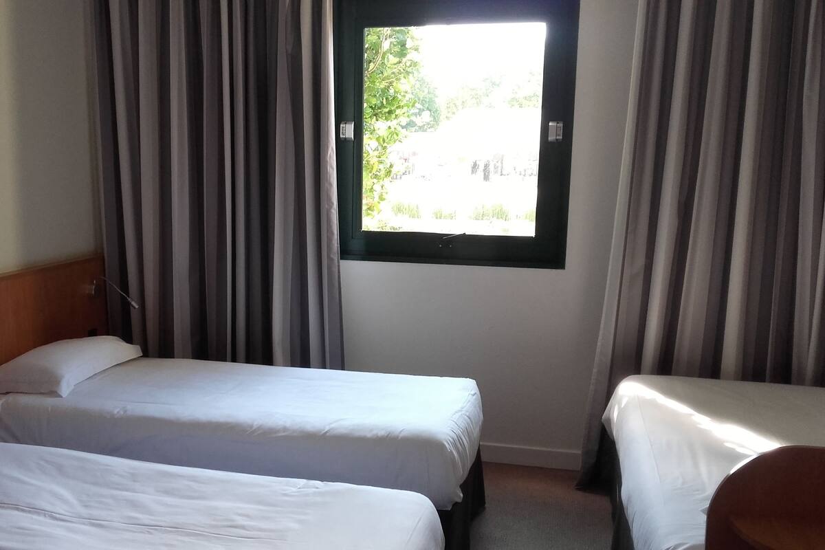 A hotel room with two single beds, a wooden headboard, a window with curtains, and a view of greenery outside.