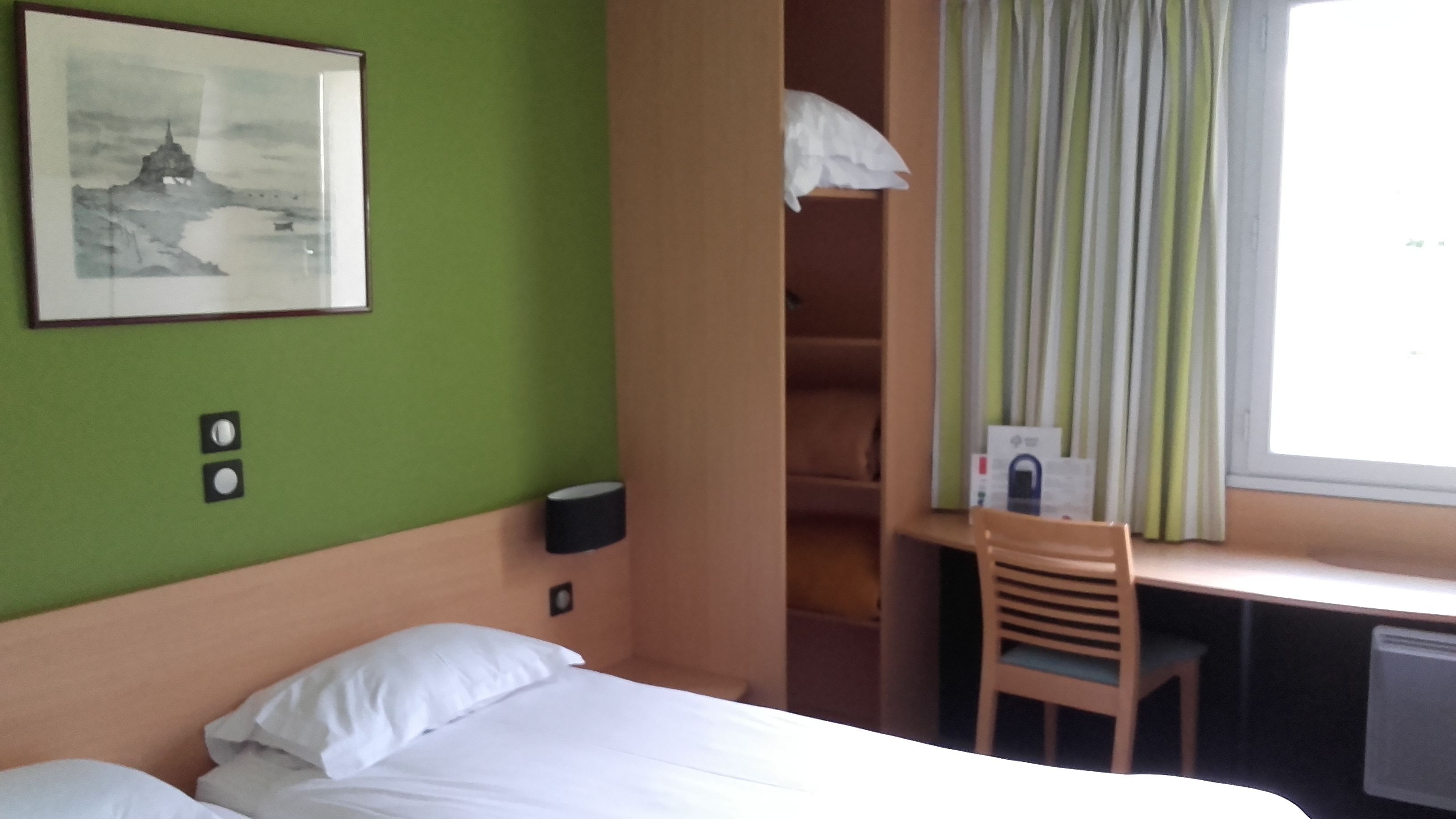 A hotel room with a bed, a desk with a chair, a window with curtains, and a wall-mounted light.