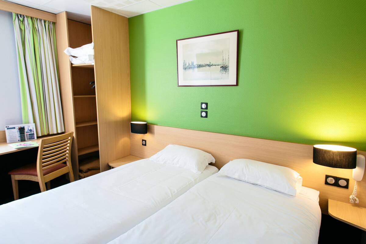 A hotel room with two beds, a desk, a chair, a wardrobe, and a painting on the wall.